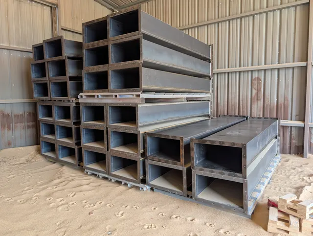 Stacked rectangular steel elevator legging sections stored in a building ready for installation